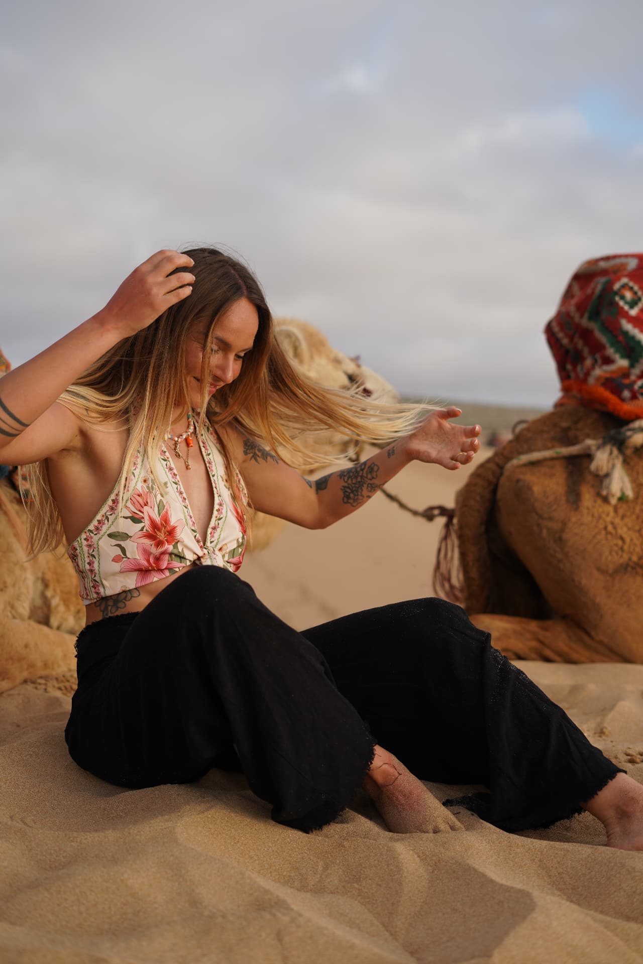 Femme assise dans le sable, cheveux au vent, dromadaires derrière elle